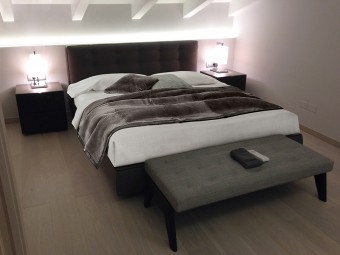 CAMERA PADRONALE. Soffitto con travi a vista. Parete dietro la testata del letto con illuminazione integrata a LED. Letto, poltrona e panca imbottiti con rivestimento in tessuto (MINOTTI). Comodini realizzati in combinazione di due materiali: Rovere Tabacco e legno laccato Moka (MINOTTI).